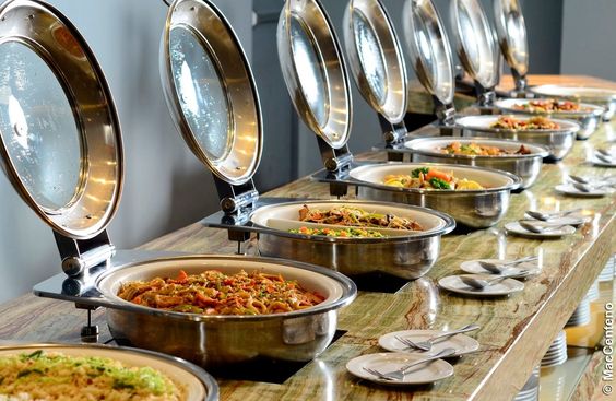 A long buffet line with multiple covered chafing dishes displaying a customized catering menu for a grand event.